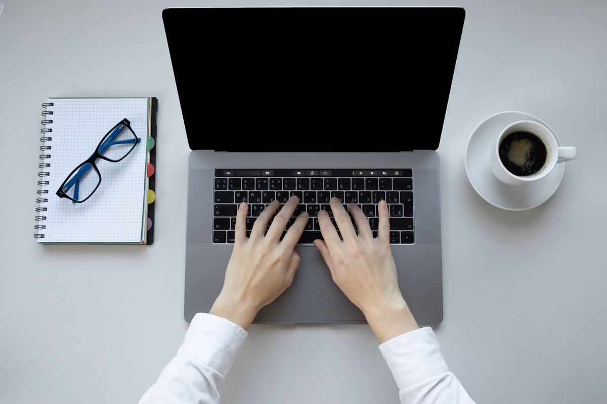 Vue du dessus : mains sur un ordinateur portable, carnet, lunettes et café sur un bureau blanc